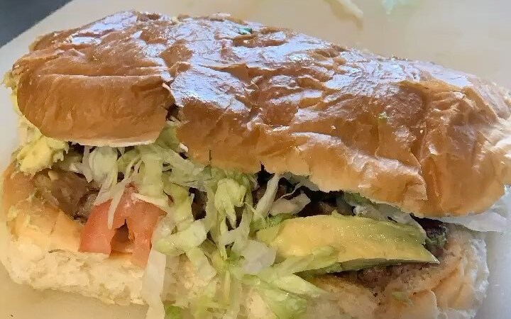 Una torta en pan dorado, partida: carne, lechuga, tomate y aguacate